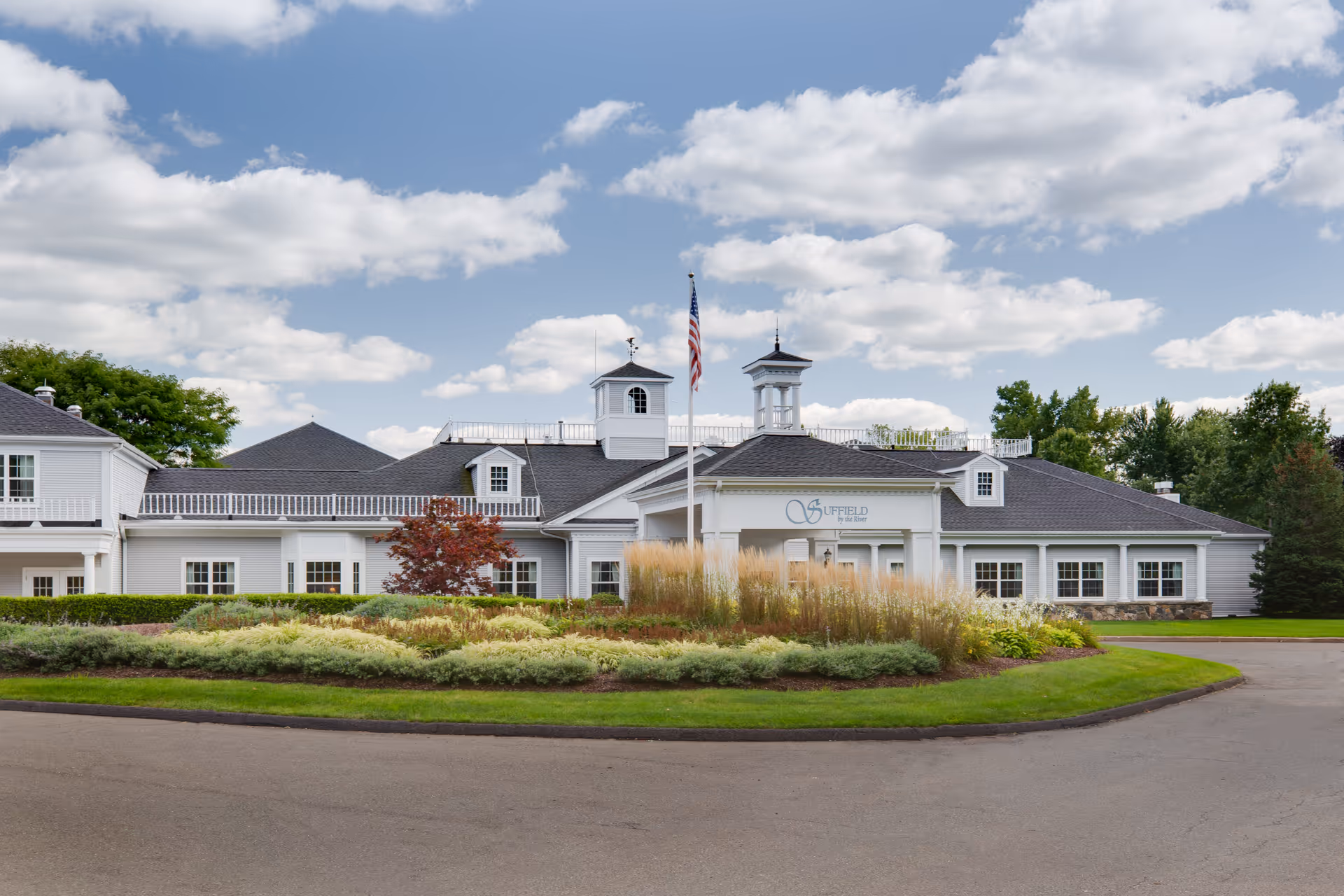Front exterior of a white senior living building with a circular driveway, landscaped island, and an American flag.