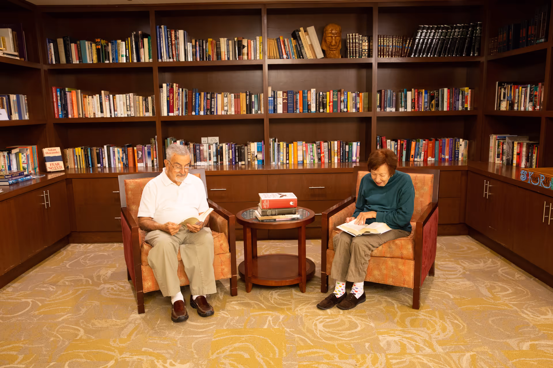Two elderly individuals sitting in armchairs in a library or reading room, each reading a book. Behind them is a large wooden bookshelf filled with numerous books and a wooden sculpture. A small round table with a few stacked books is placed between them.