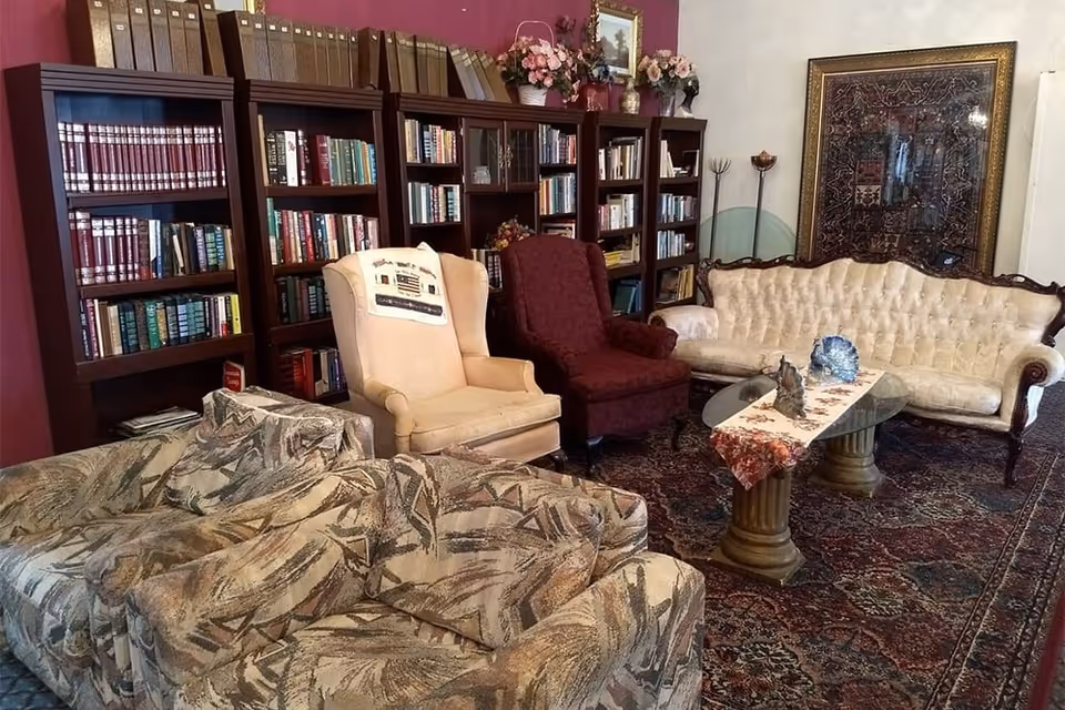 A cozy living room area with a patterned sofa, a beige wingback chair with a decorative throw, a dark red armchair, and a cream tufted sofa. A glass coffee table with a decorative runner and figurines sits on a large patterned rug. Behind the seating area is a wall with multiple wooden bookshelves filled with books and decorative items, and a large framed tapestry hangs on the adjacent wall.