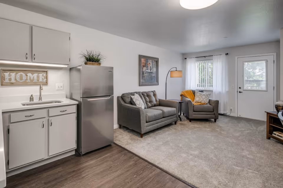 A cozy living area in a retirement community featuring a small kitchen with white cabinets, a stainless steel refrigerator, and a sink. Adjacent to the kitchen is a carpeted sitting area with a gray loveseat and armchair, a floor lamp, a framed painting on the wall, and a window with sheer white curtains letting in natural light. A door with a window is visible in the background.