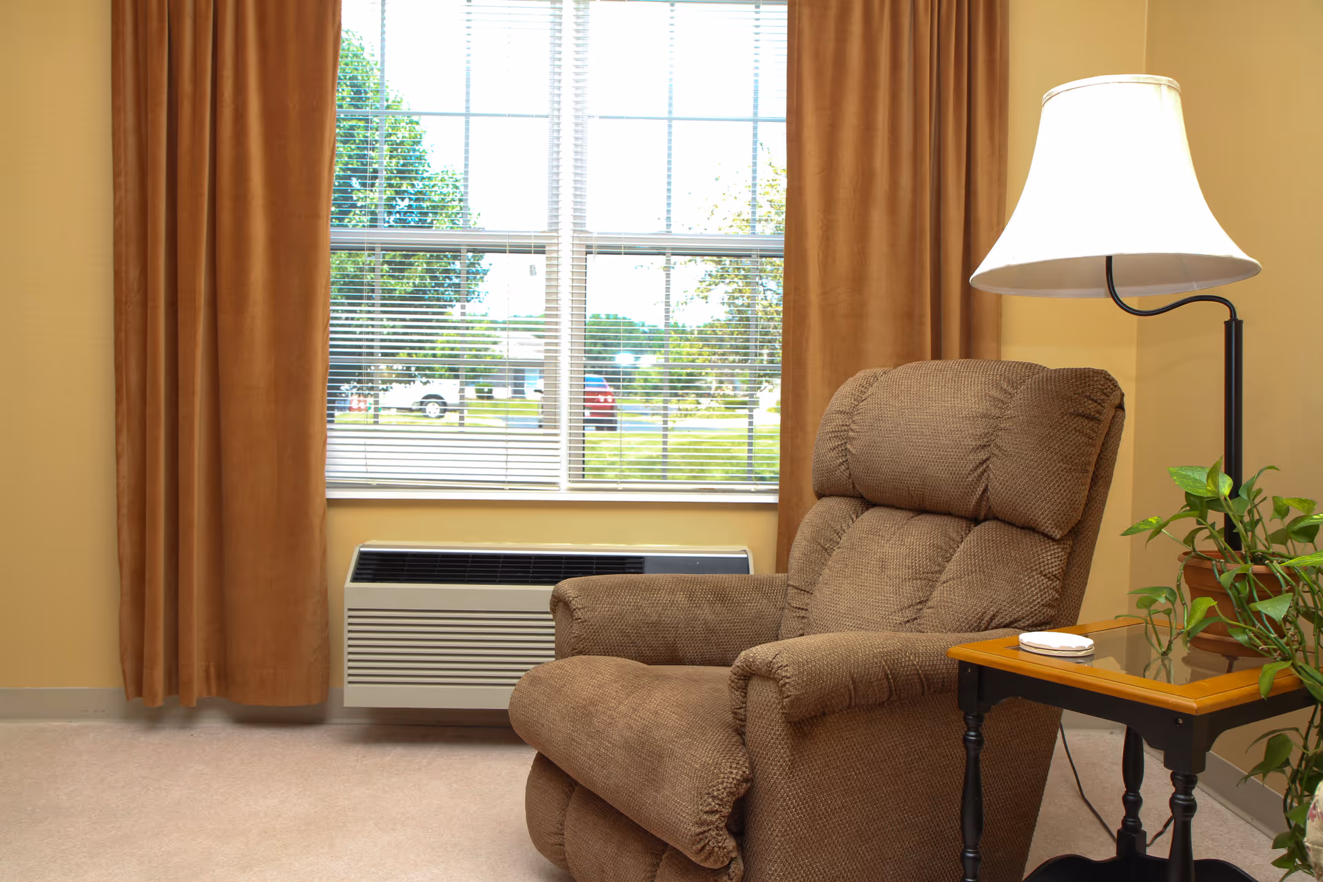A cozy corner of a room featuring a brown upholstered recliner chair next to a wooden side table with a potted plant and a white lampshade on a black lamp. Behind the chair is a large window with closed blinds and brown curtains, showing a view of trees and parked cars outside.