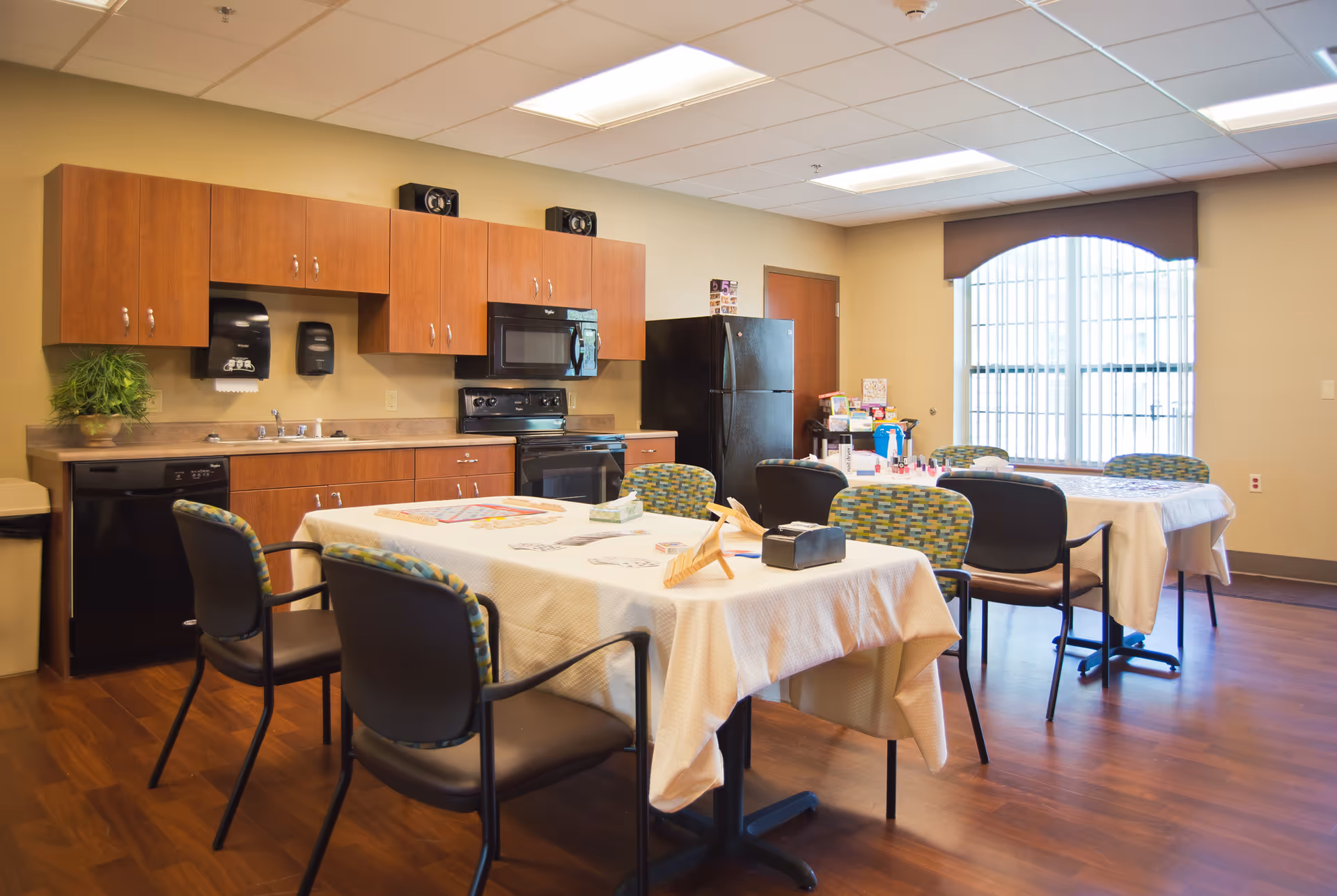 Interior view of a communal kitchen and dining area with wooden cabinets, black appliances including a refrigerator, stove, microwave, and dishwasher. Two tables covered with white tablecloths are surrounded by chairs, and various items such as board games and nail polish are placed on the tables. A large window with vertical blinds allows natural light into the room.