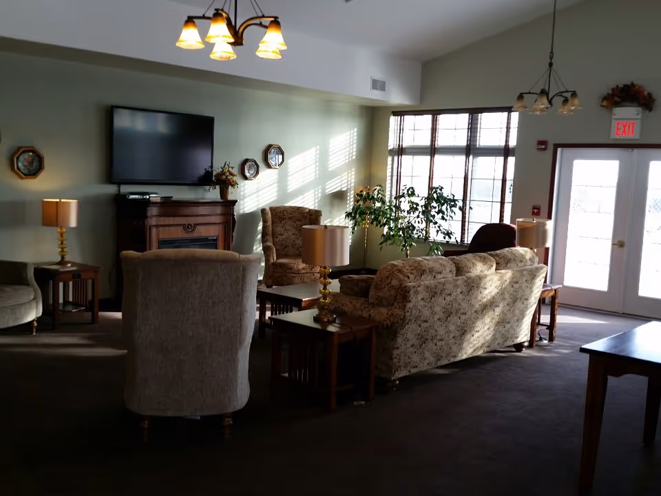 A cozy living room area with floral patterned sofas and armchairs arranged around wooden side tables with lamps. A flat-screen TV is mounted on the wall above a wooden fireplace. Large windows with blinds allow natural light to fill the room. The room has soft green walls and carpeted flooring, with ceiling light fixtures and an exit door visible in the background.