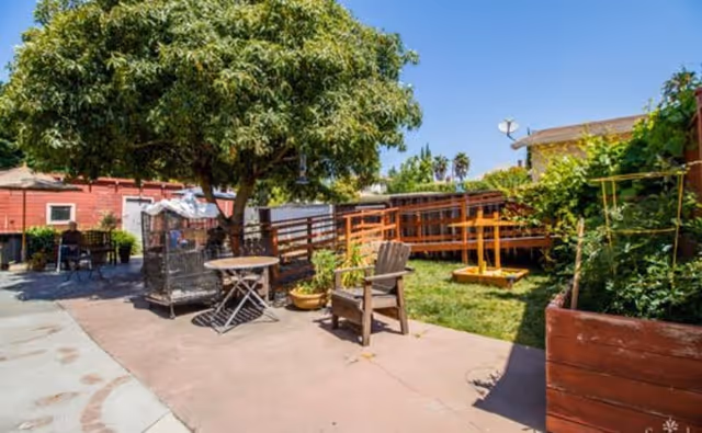 Outdoor patio area with a large tree providing shade, a round table with chairs, a wooden chair, potted plants, a wooden ramp, and a swing set in a grassy area under a clear blue sky.