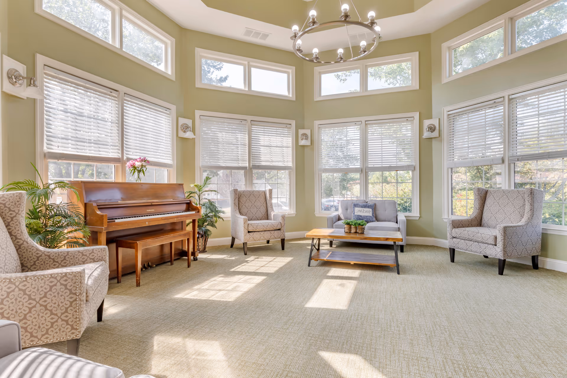 Bright communal sitting room with large windows, a piano, sofa, armchairs, and a central coffee table.