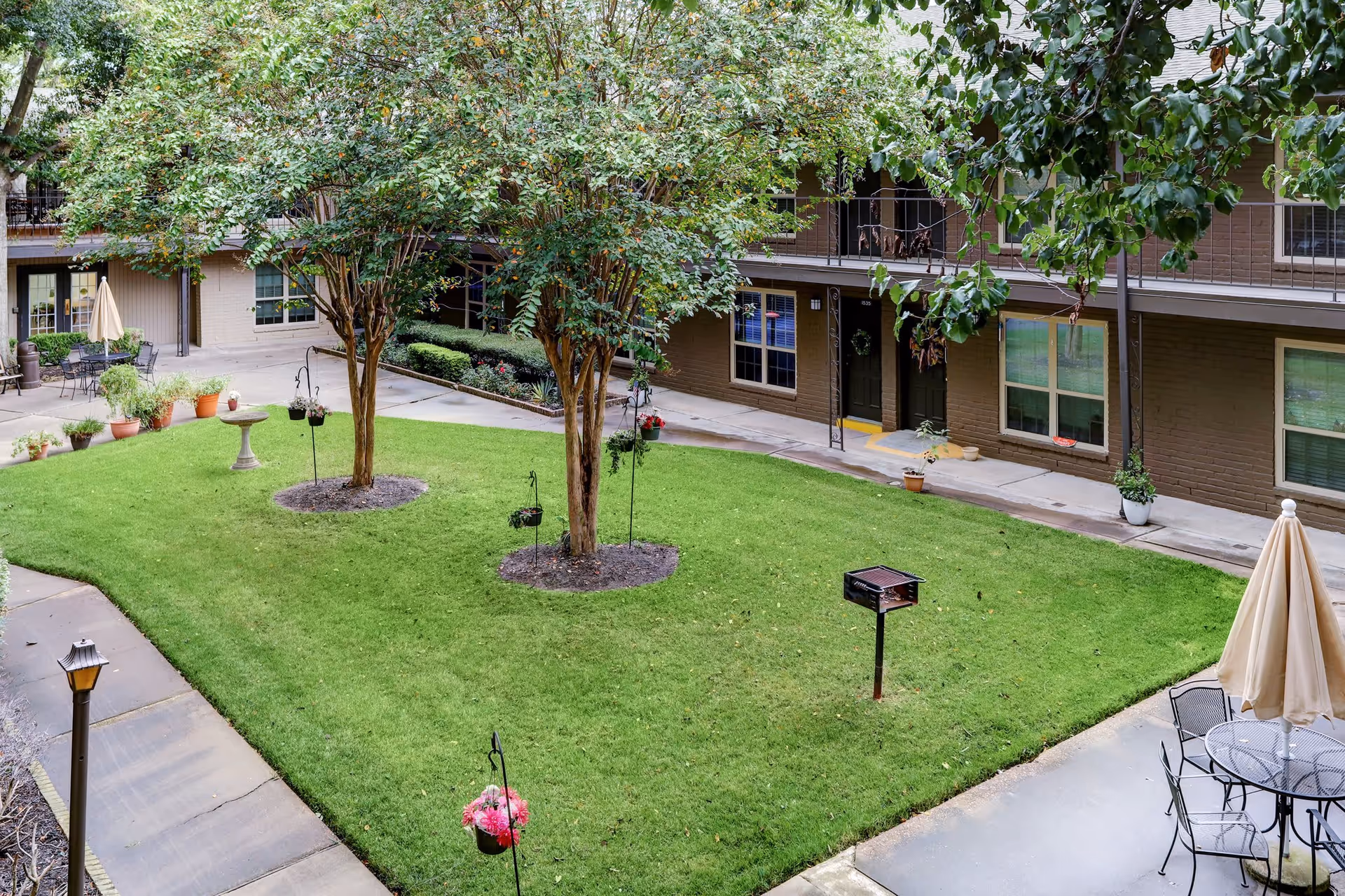 A well-maintained outdoor courtyard area with green grass, two trees, hanging flower pots, a birdbath, a barbecue grill, and patio furniture with an umbrella. Surrounding the courtyard are two-story brown brick buildings with windows and doors, some decorated with plants and wreaths.