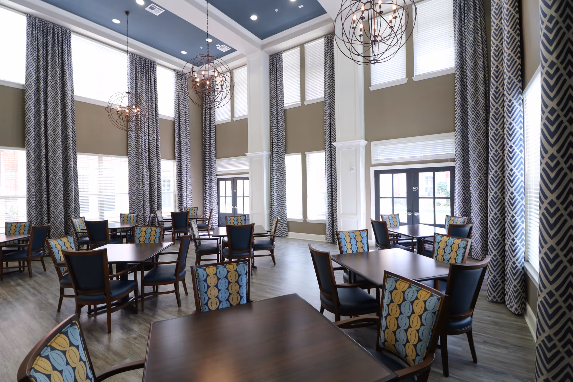 A spacious dining room with multiple dark wood tables and chairs featuring patterned upholstery. The room has tall windows with long patterned curtains, light-colored walls, and modern spherical pendant lights hanging from a high ceiling.