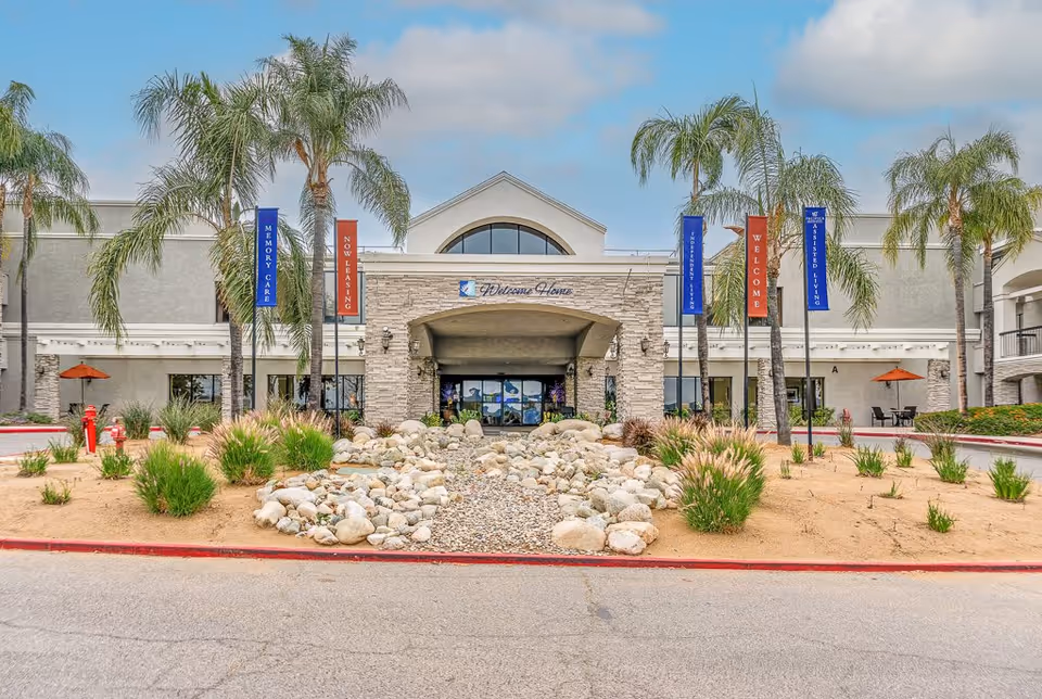 Front exterior view of Menifee Senior Living facility with palm trees, landscaped rocks, and banners indicating memory care, now leasing, independent living, welcome, and assisted living services.