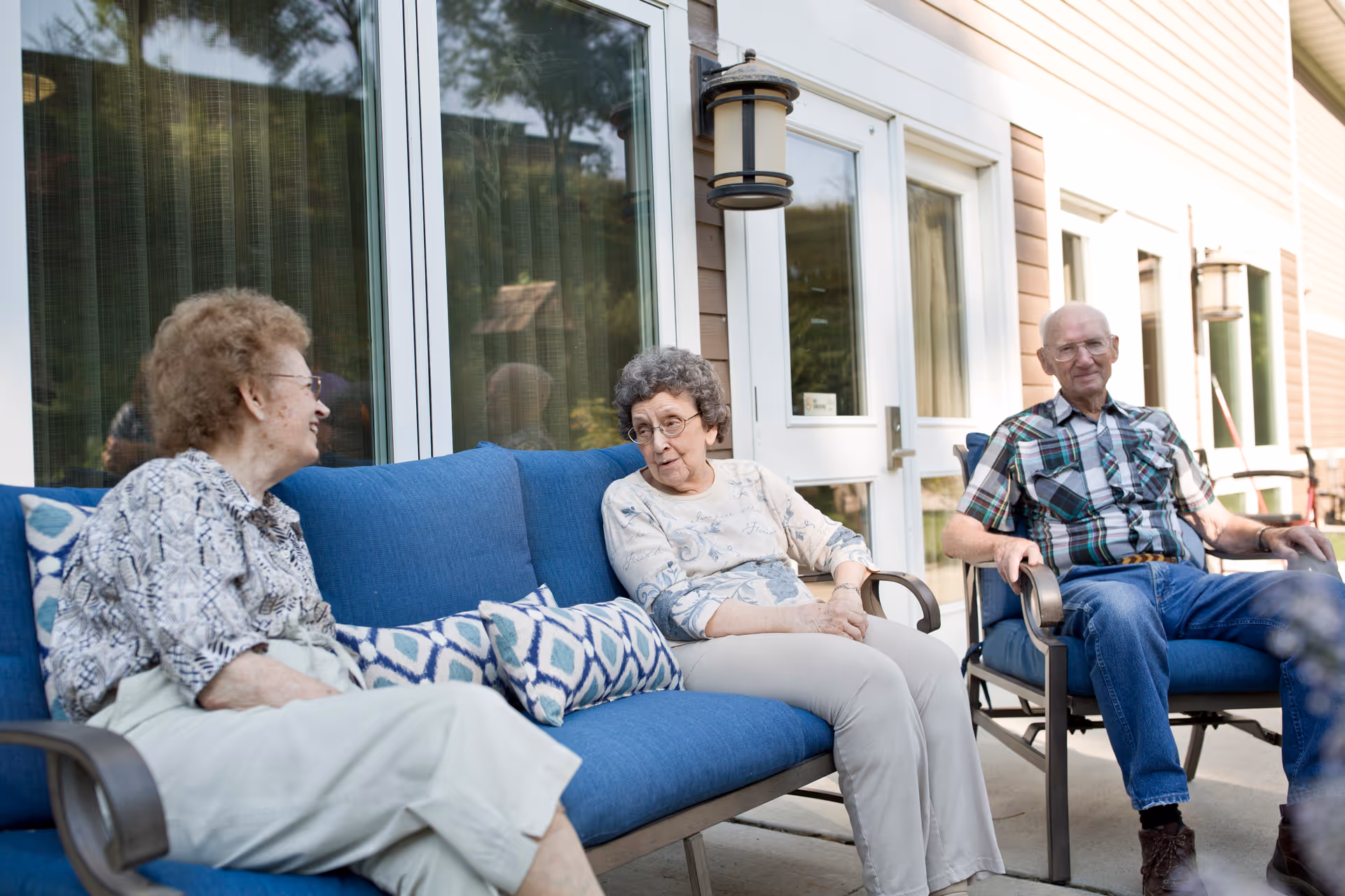 Three elderly people sitting and chatting on blue cushioned outdoor chairs and sofa on a patio outside a building with large windows and a door.
