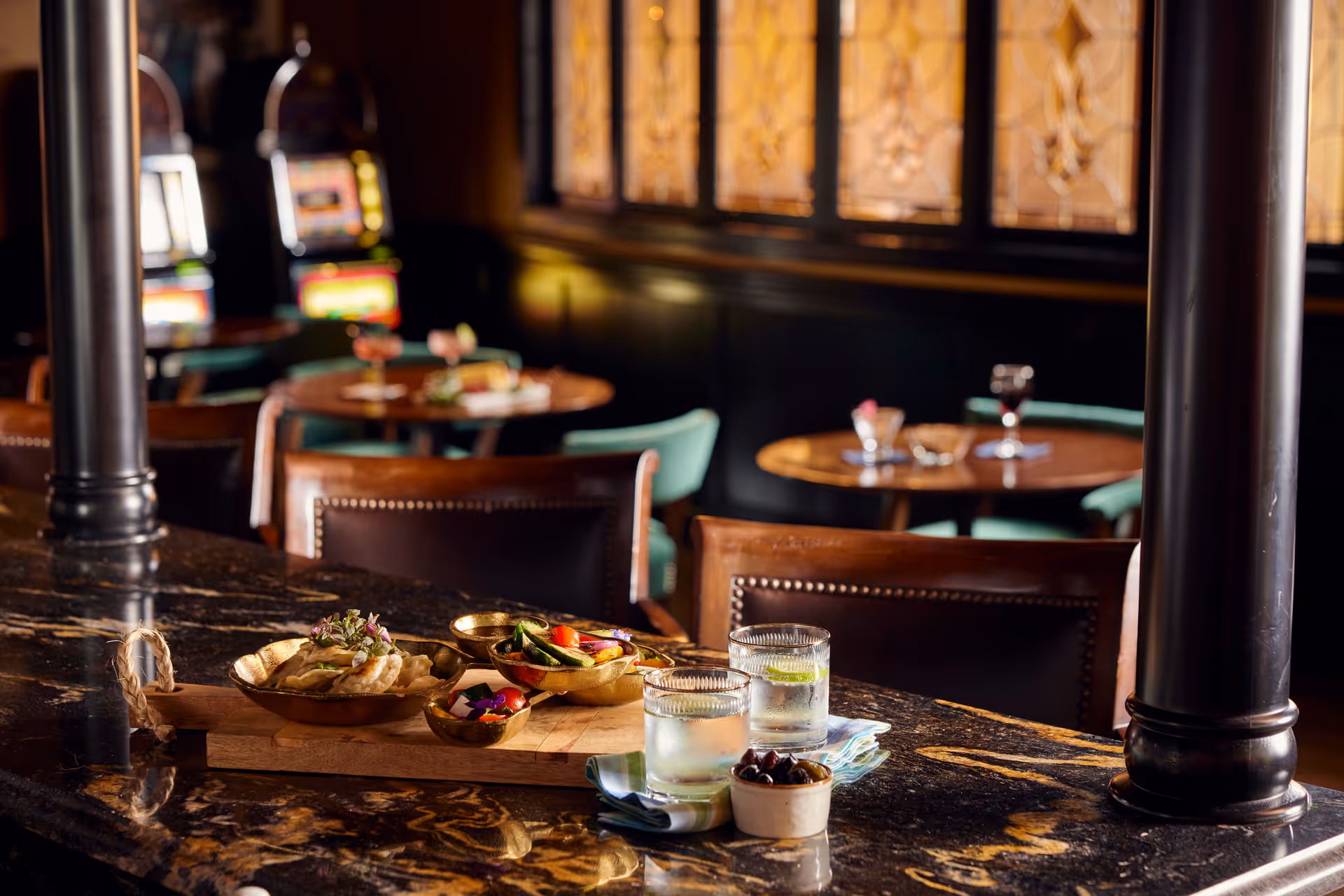 A close-up view of a dark marble countertop with a wooden tray holding small bowls of food and two glasses of water with lime. In the background, there are round tables with chairs and stained glass windows, creating a cozy dining atmosphere.
