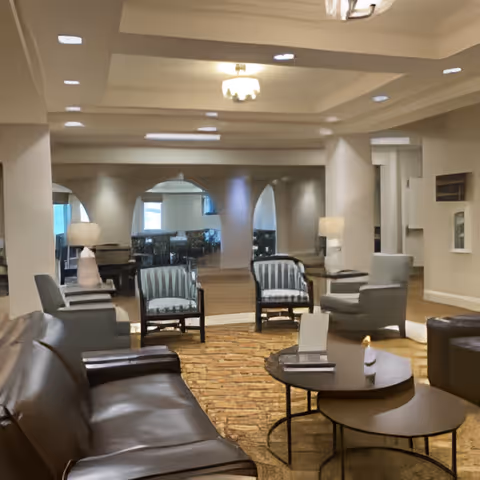 A spacious and well-lit living room area with a brown leather sofa, two striped armchairs, and two gray armchairs arranged around a set of round nesting coffee tables. The room features a patterned carpet, modern ceiling lights, and lamps on side tables. The walls have large mirrors and archways leading to other parts of the facility.