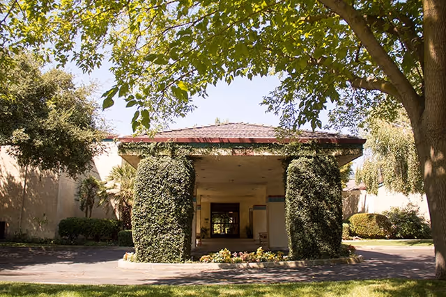 Entrance of a building with a covered driveway supported by two large columns covered in greenery, surrounded by trees and bushes under a clear sky.