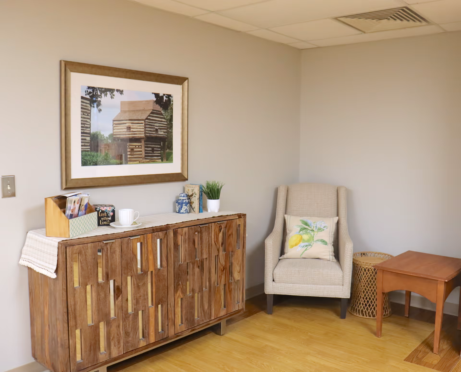 A cozy corner of a room featuring a wooden cabinet with decorative items on top, including a small plant, a cup and saucer, and a framed picture of a rustic wooden building hanging on the wall above. Next to the cabinet is a beige upholstered armchair with a decorative pillow, a small wooden side table, and a wicker basket on a light wood floor.