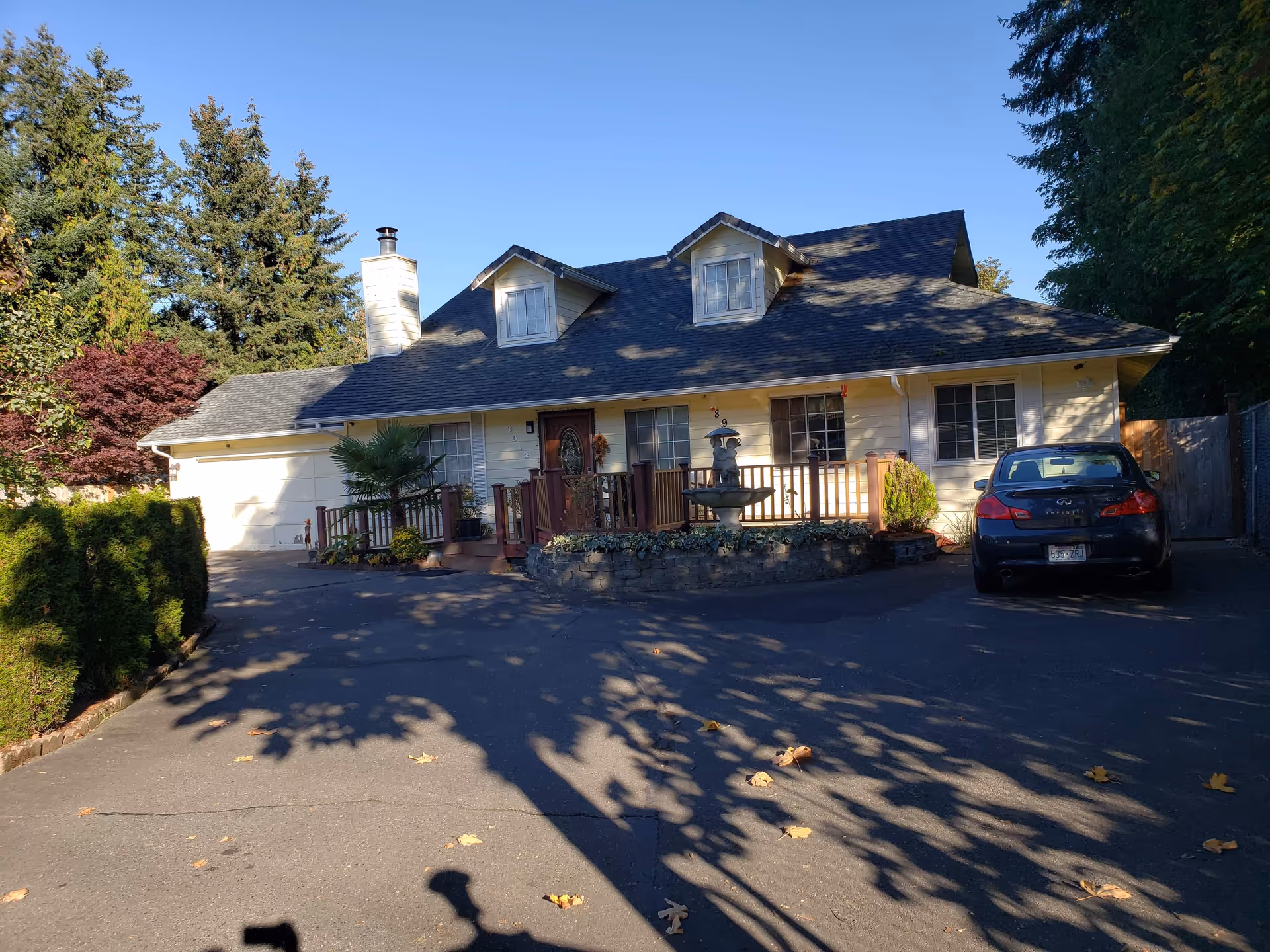 A suburban house front with dormer windows, a wooden porch and a driveway leading to a parked car.