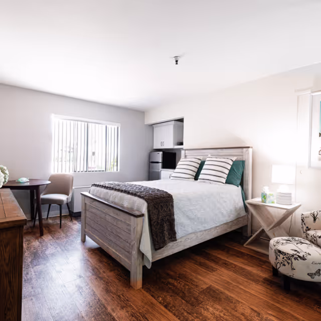 A bright and clean bedroom with a wooden bed frame, white bedding, and striped pillows. The room features wooden flooring, a small round table with a chair near a window with vertical blinds, a nightstand with a lamp and tissue box, a patterned armchair, and a kitchenette area with a microwave and mini fridge.