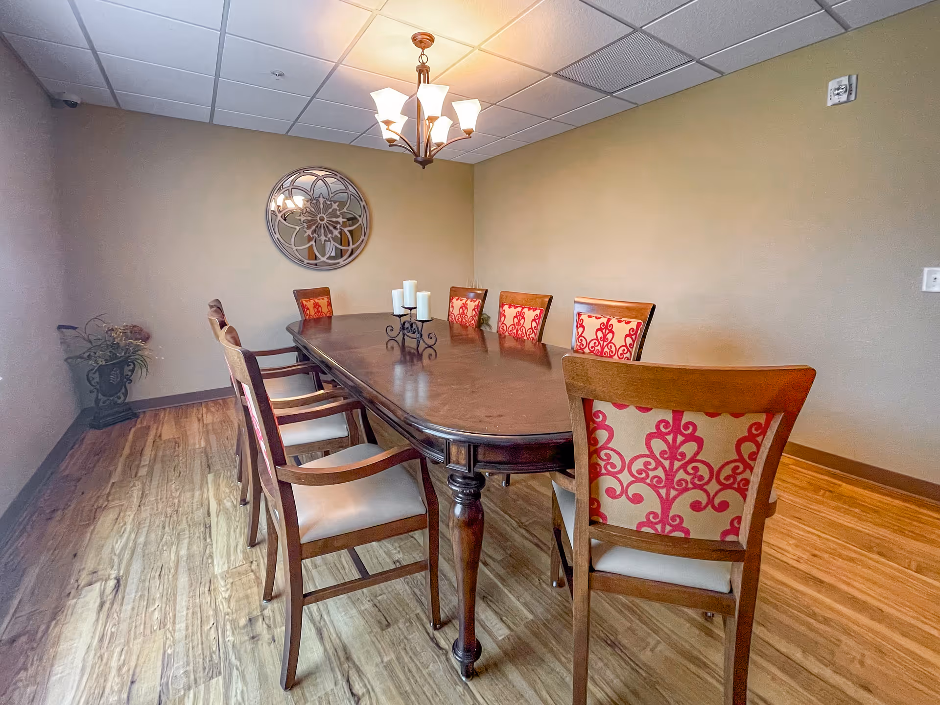 A dining room with a long wooden table surrounded by eight chairs, each with red and beige patterned upholstery on the backrest. A decorative round mirror hangs on the beige wall, and a chandelier with multiple lights is suspended from the ceiling. There is a small plant in a decorative pot in the corner of the room, and the floor is made of wood.