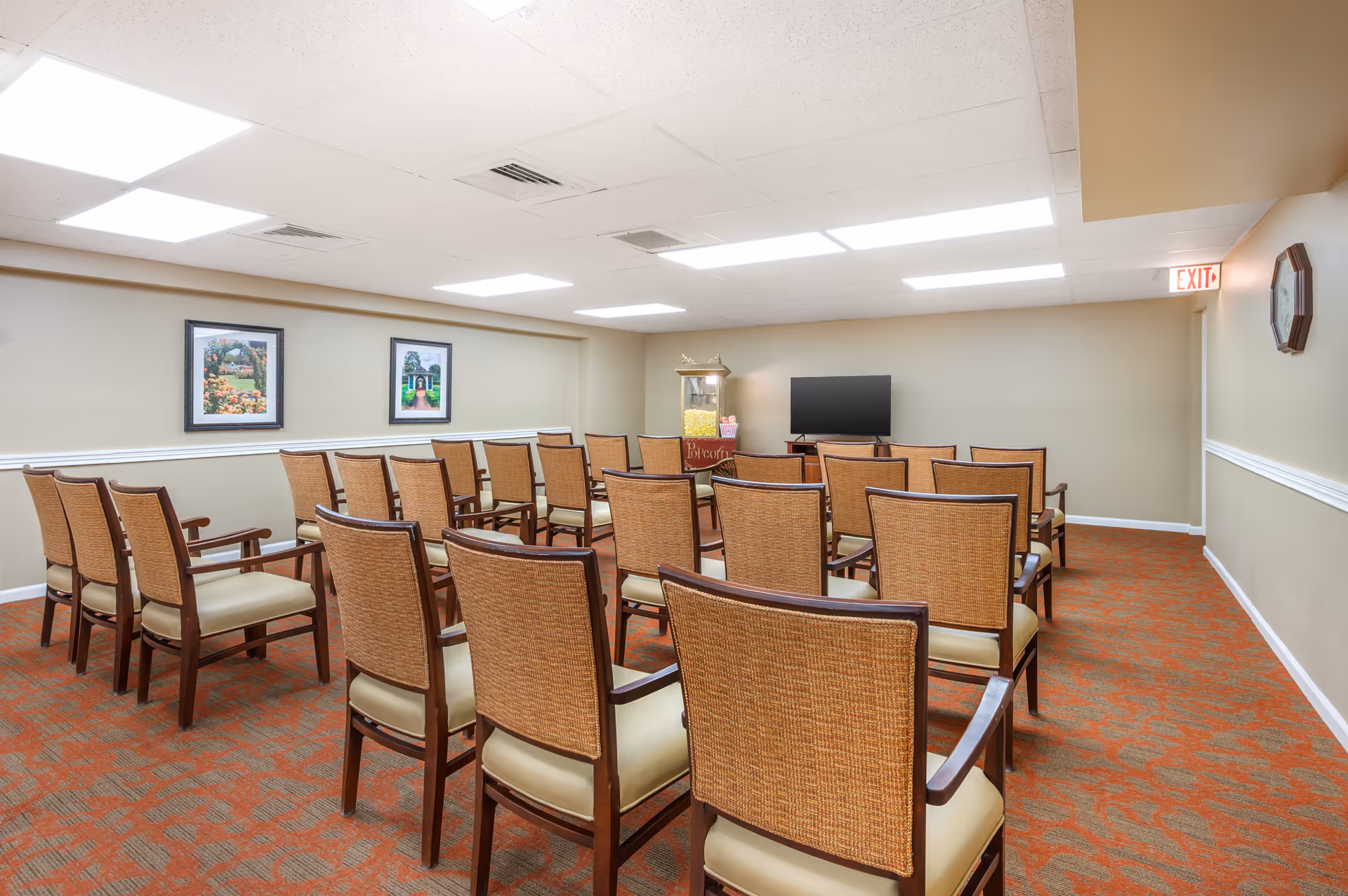 Rows of wooden chairs arranged facing a TV and popcorn machine in a bright activity/meeting room with framed pictures on the wall.