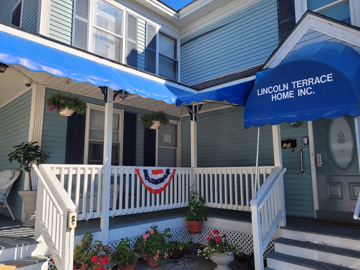 Front porch of a light blue building with white railings and steps. A blue awning above the entrance reads 'LINCOLN TERRACE HOME INC.'. There are hanging flower pots and potted plants decorating the porch area, along with a red, white, and blue bunting on the railing.