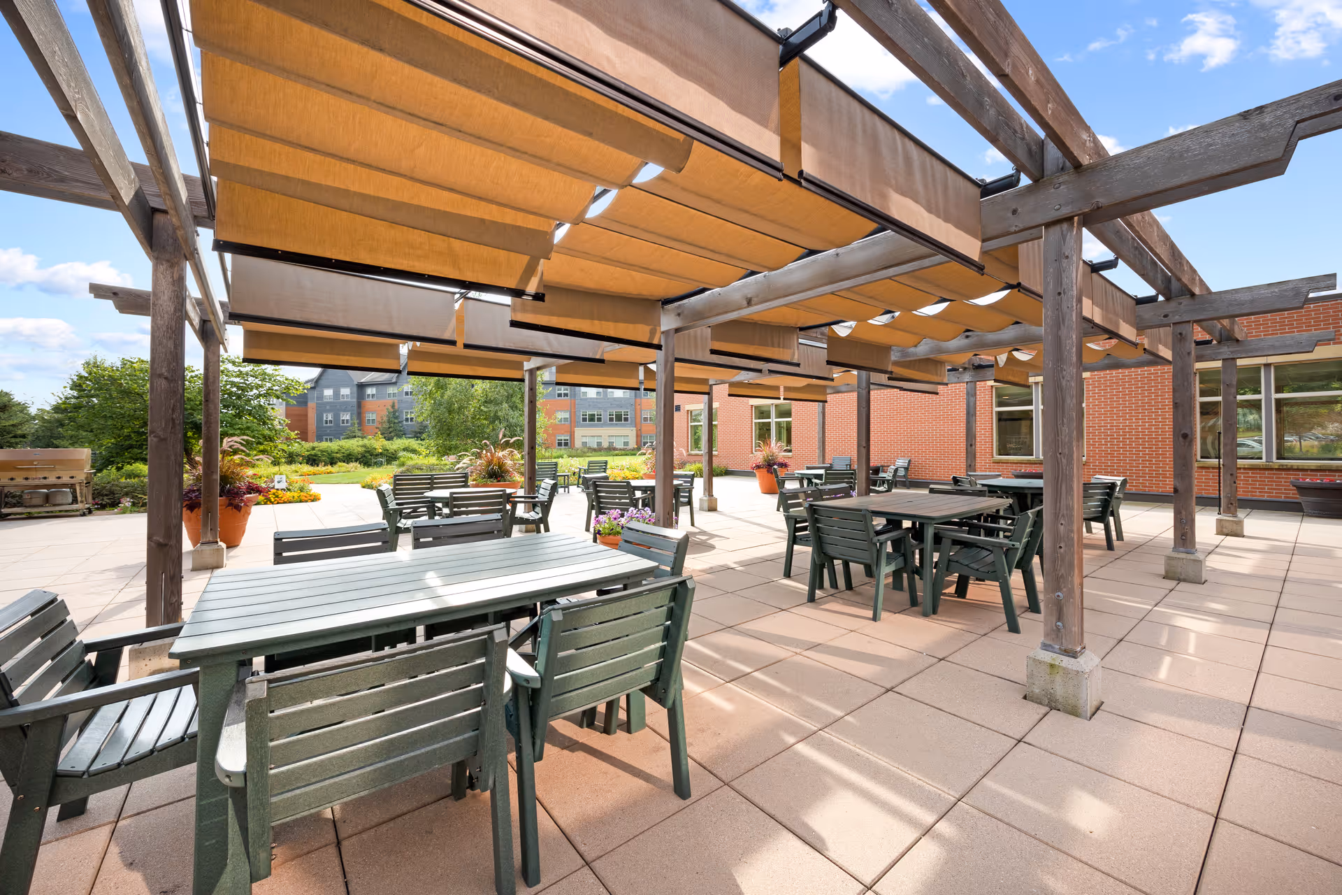 Outdoor patio with a wooden pergola and fabric shades, several dining tables and chairs, and a brick building and garden in the background.
