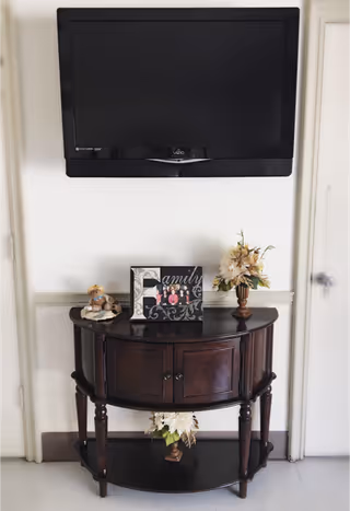 Wall-mounted flat-screen TV above a dark wooden console table holding a framed 'Family' photo, floral arrangements, and decorative figurines in an interior hallway.