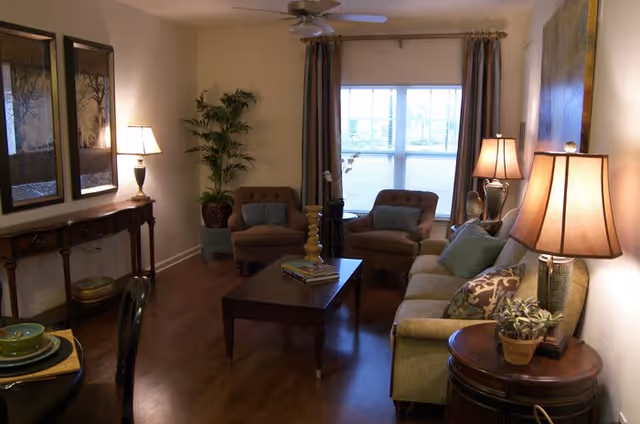 A cozy living room with a sofa, two armchairs, a coffee table with books and decorative items, a side table with a lamp and a potted plant, a console table with two lamps and framed artwork above it, and a window with curtains letting in natural light.