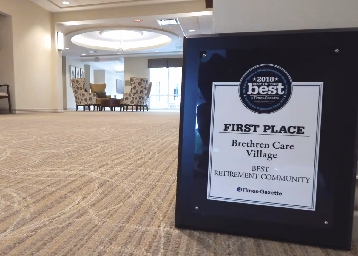 A framed award plaque placed on a carpeted floor in a lounge area with chairs and a table in the background. The plaque reads '2018 Best of the Best, First Place, Brethren Care Village, Best Retirement Community, Times-Gazette.'