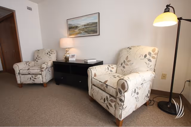 A cozy interior seating area with two beige armchairs featuring a leaf pattern, a black console table between them holding a lamp, a small framed photo, and a book. A floor lamp with a yellow shade stands to the right, and a landscape painting hangs on the white wall above the console table.