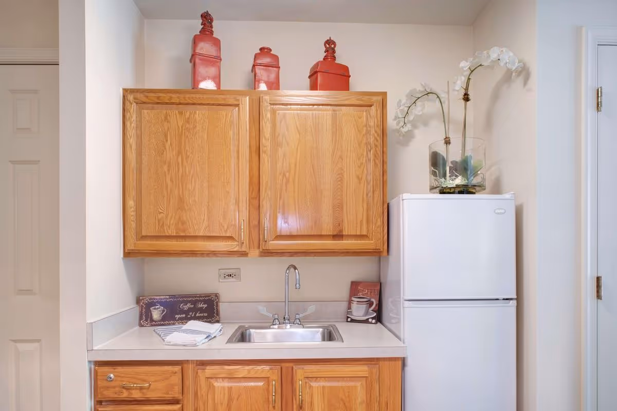 Small kitchenette with oak cabinets above a countertop sink and a white refrigerator with decorative items on top.