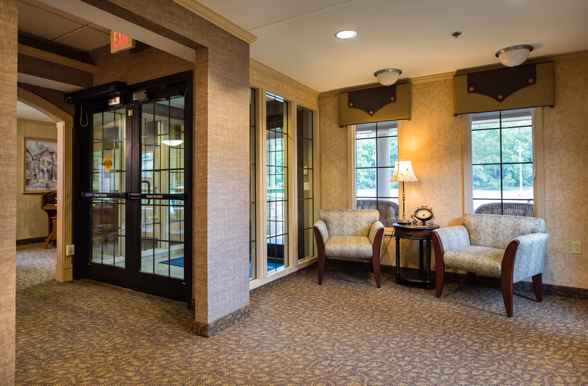 A seating area in an assisted living facility with two upholstered armchairs around a side table and lamp, windows overlooking greenery, and glass double entrance doors.