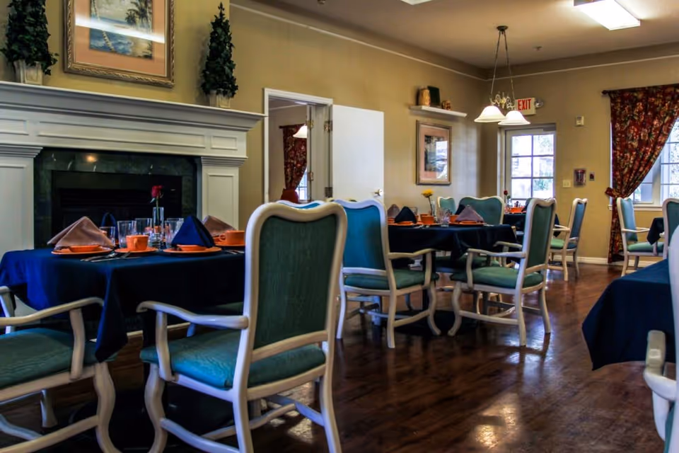 Dining room with blue-tablecloth tables set for meals and green upholstered chairs arranged around a fireplace.