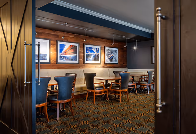 Interior view of a dining area with wooden tables and chairs, a cushioned bench along the wall, and framed nautical-themed photographs hanging on a wooden accent wall. The room features patterned carpet flooring and is partially visible through open double doors with metal handles.