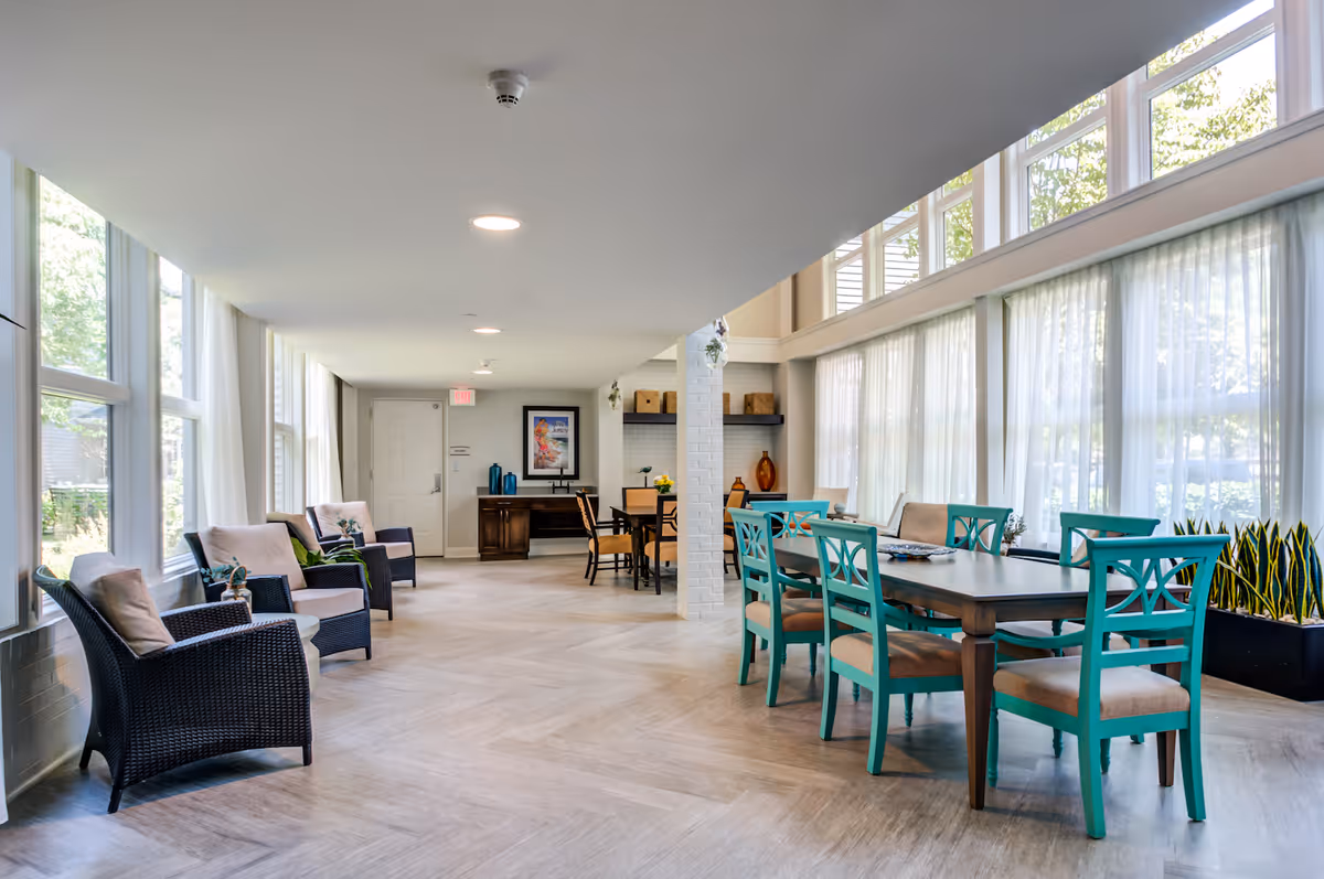 A bright and spacious common area with large windows covered by sheer white curtains. The room features a row of black wicker chairs with beige cushions on the left side and a dark wooden table with turquoise chairs on the right. There is a white brick column in the center and a wooden desk with decorative items and a framed picture on the far wall.