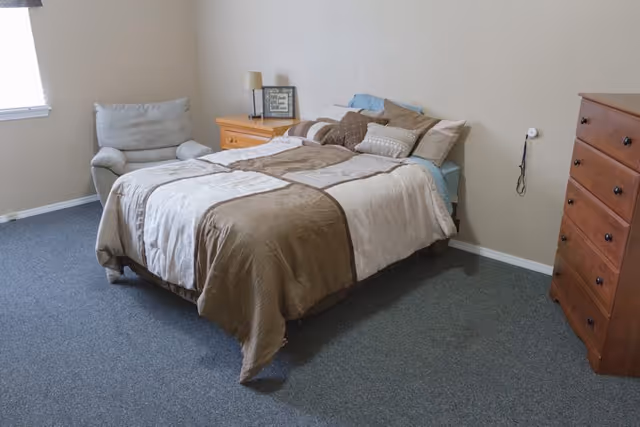 Furnished bedroom with a made bed, armchair by the window, nightstand with lamp, and a tall wooden dresser.