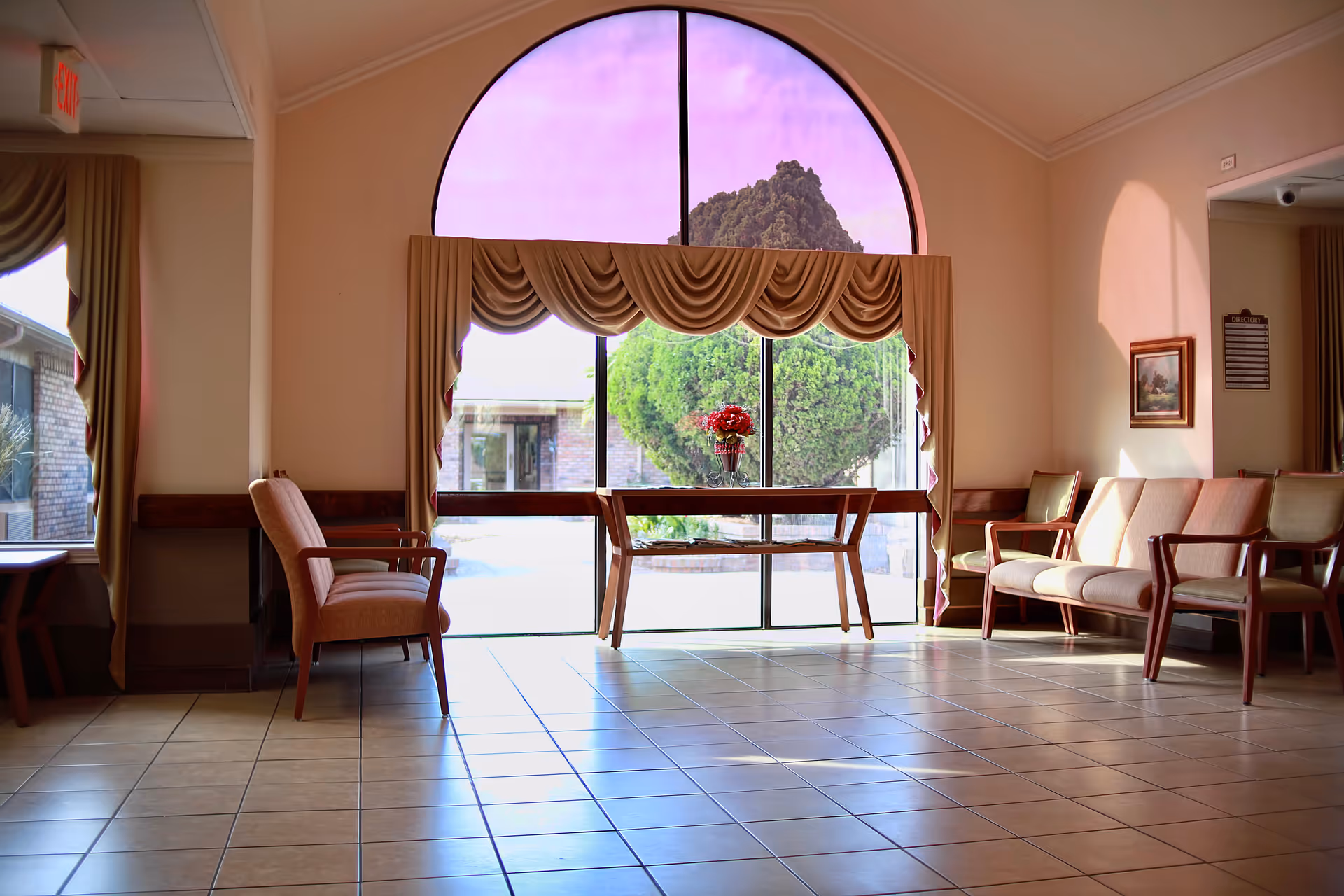 Sunlit seating area with chairs and a table holding a vase of flowers in front of a large arched window.