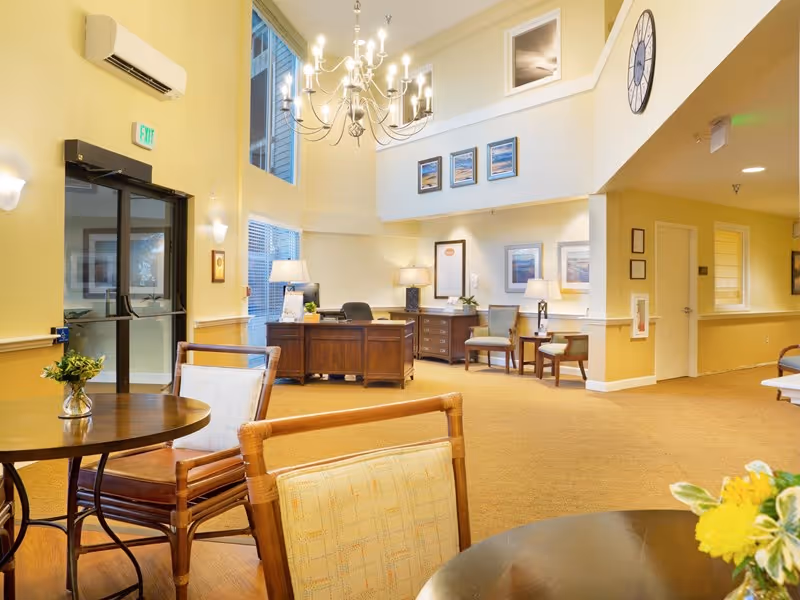 Bright two-story assisted living lobby with seating areas, a reception desk, chandelier, and tables.