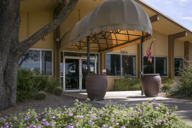 Front entrance of a rehabilitation center with a canopy over glass double doors, large planters, an American flag, and landscaped foreground.