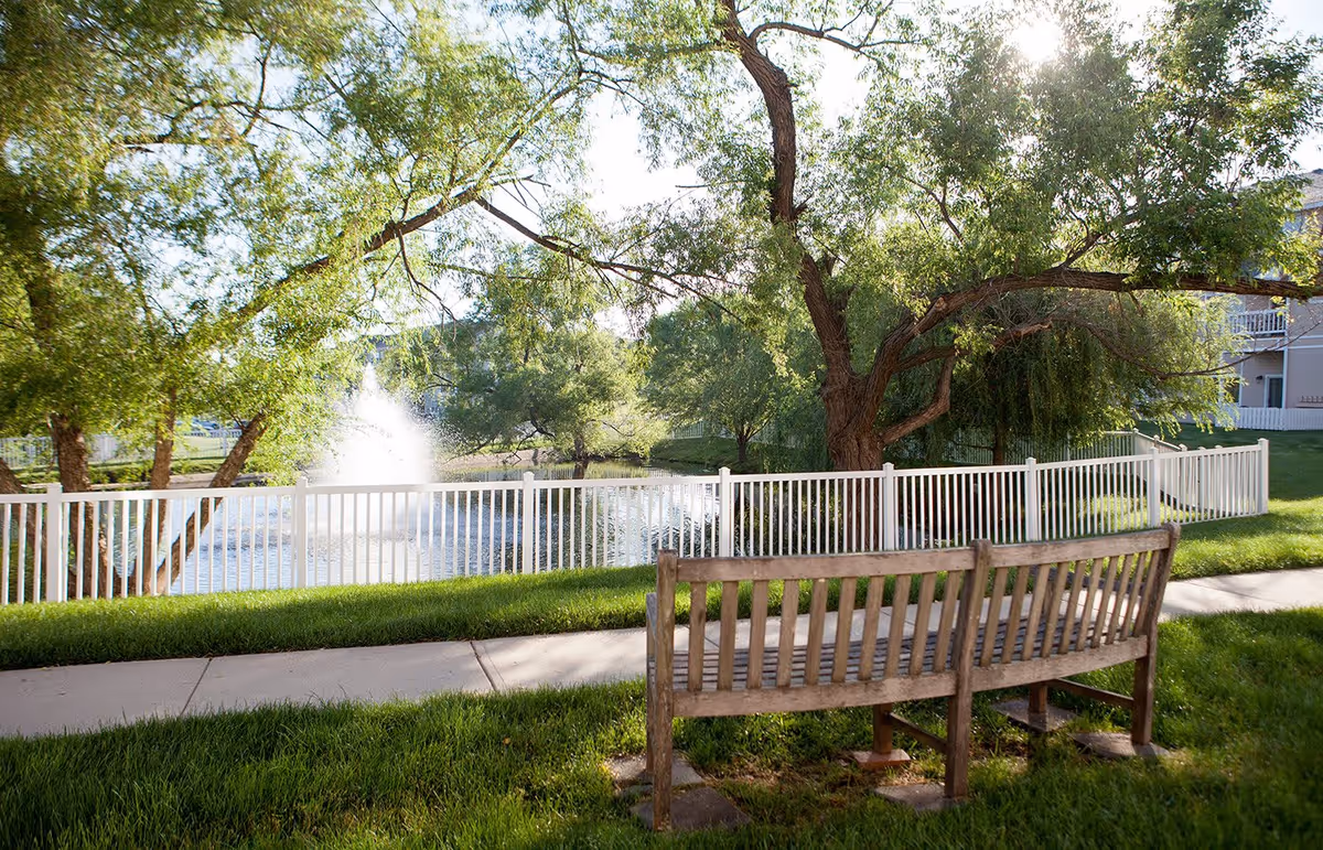 A peaceful outdoor scene featuring a wooden bench on a grassy area next to a paved walkway. Behind the bench is a white fence enclosing a pond with a water fountain spraying water into the air. Large leafy trees provide shade and sunlight filters through the branches.