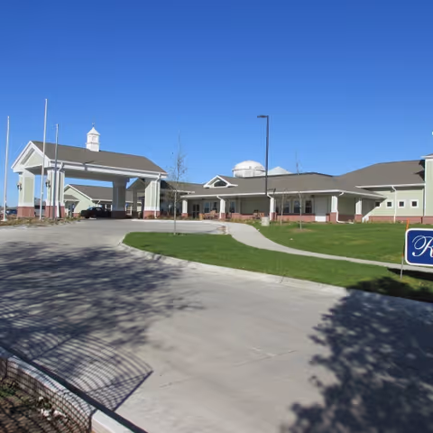 Front entrance and porte-cochere of a single-story senior care building with a driveway and green lawn under a clear blue sky.
