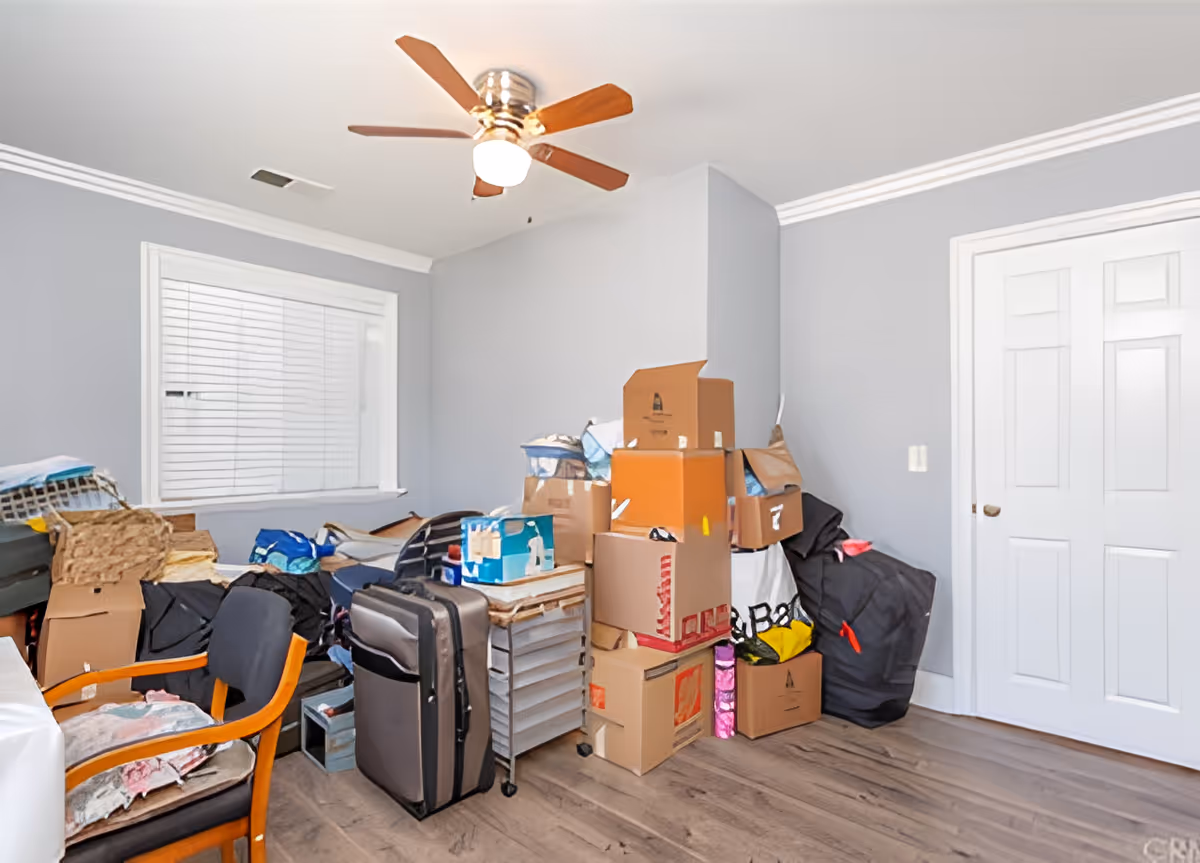 A cluttered room with a ceiling fan, gray walls, a window with blinds, a closed white double door, and a pile of packed cardboard boxes, suitcases, and bags stacked against the wall. There is also a wooden chair with a cushion and some folded fabric on it.
