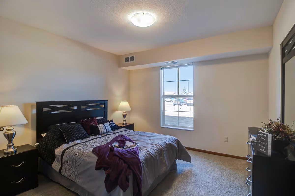 A bedroom with a large bed featuring a dark wooden headboard, patterned bedding, and several pillows. There are two bedside tables with lamps on each side of the bed. A window with a view of parked cars outside lets in natural light. A dresser with a mirror and decorative items is on the right side of the room. The walls are painted beige and the floor is carpeted.