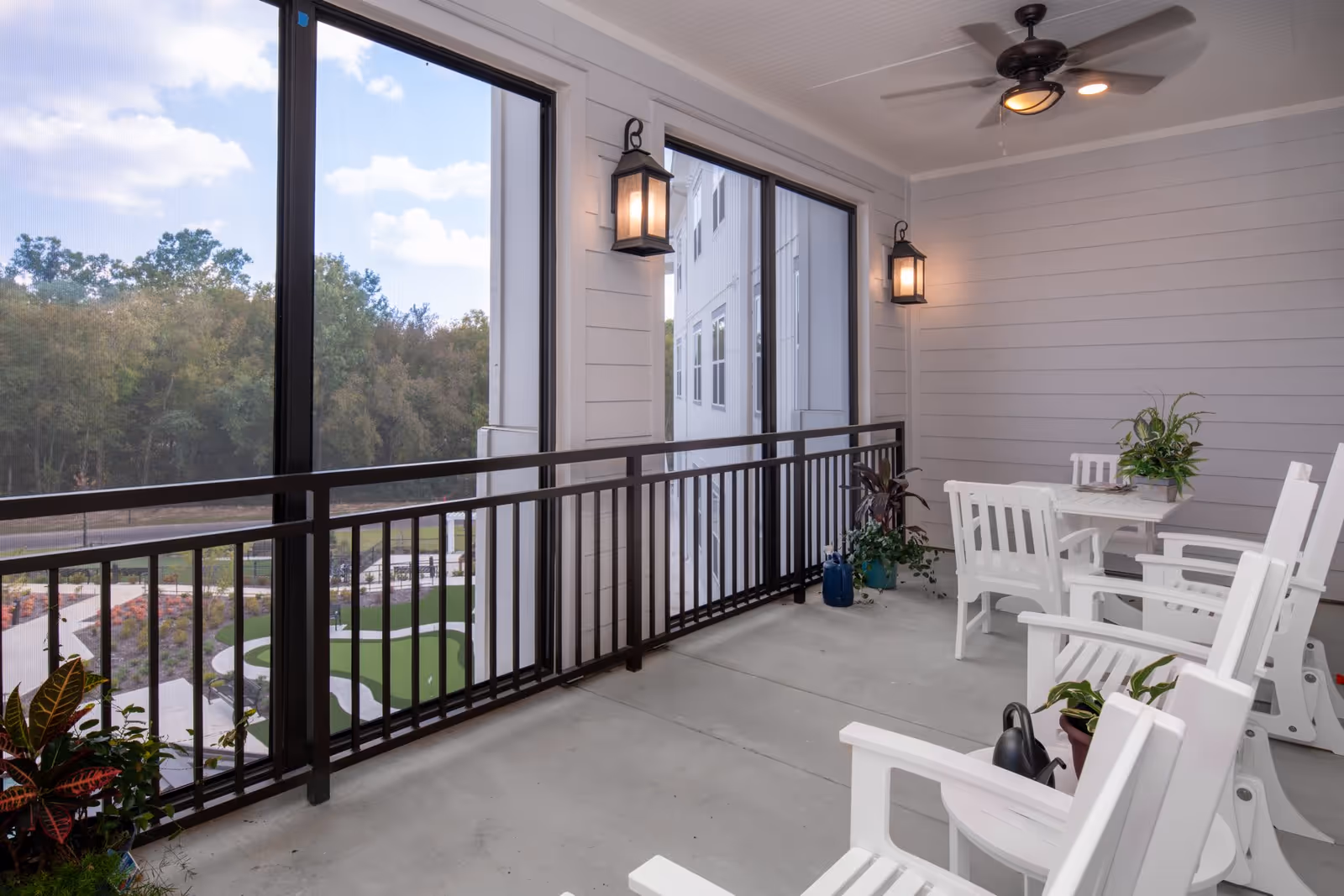 A screened-in balcony with white wooden chairs and a small table with a plant on it. The balcony overlooks a landscaped outdoor area with trees and a putting green. The walls are light gray with two lantern-style wall lights and a ceiling fan above.