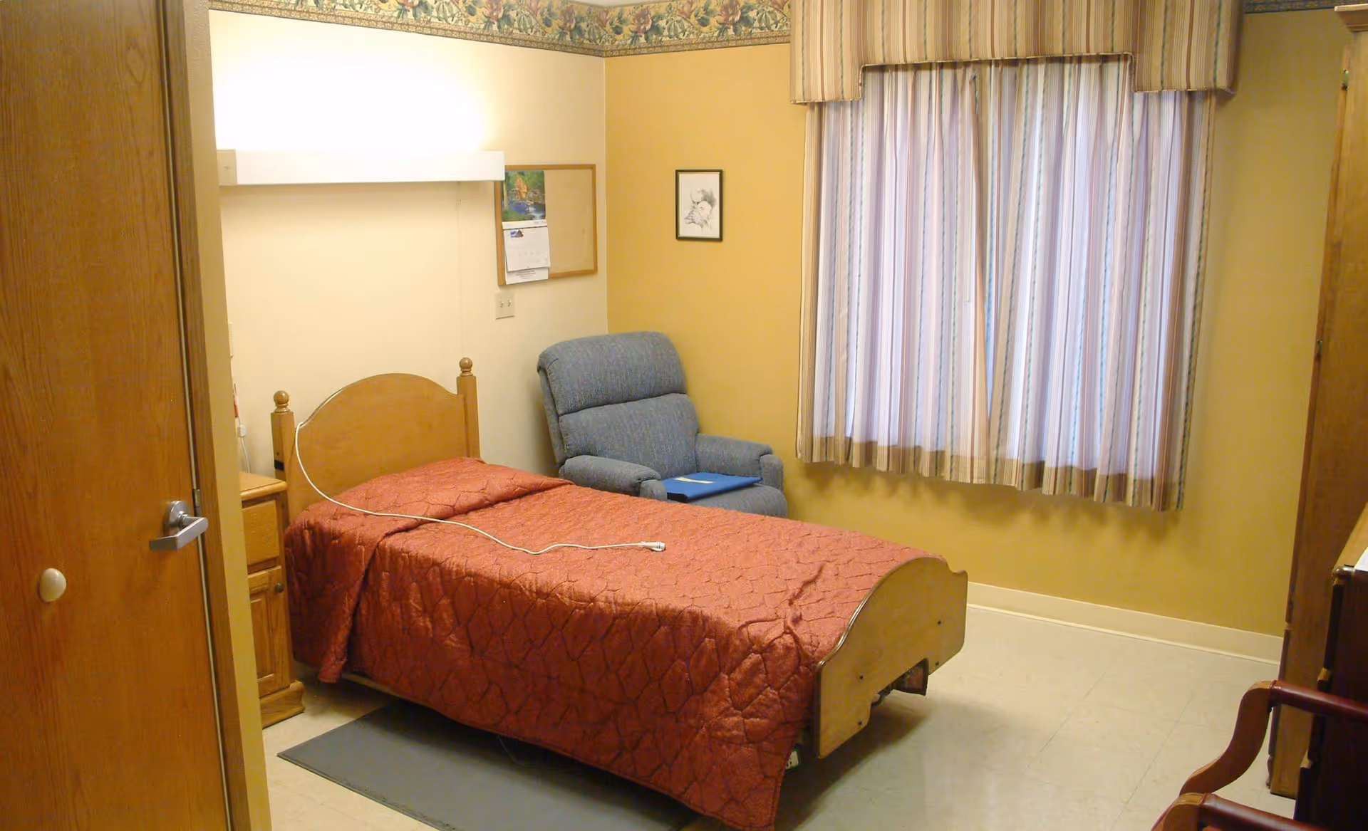 A small bedroom in a senior care facility with a single wooden bed covered with a red quilt. Next to the bed is a wooden nightstand and a blue upholstered armchair. The room has beige walls, a window with striped curtains, and a wooden door partially open. A calendar and a framed picture hang on the wall.