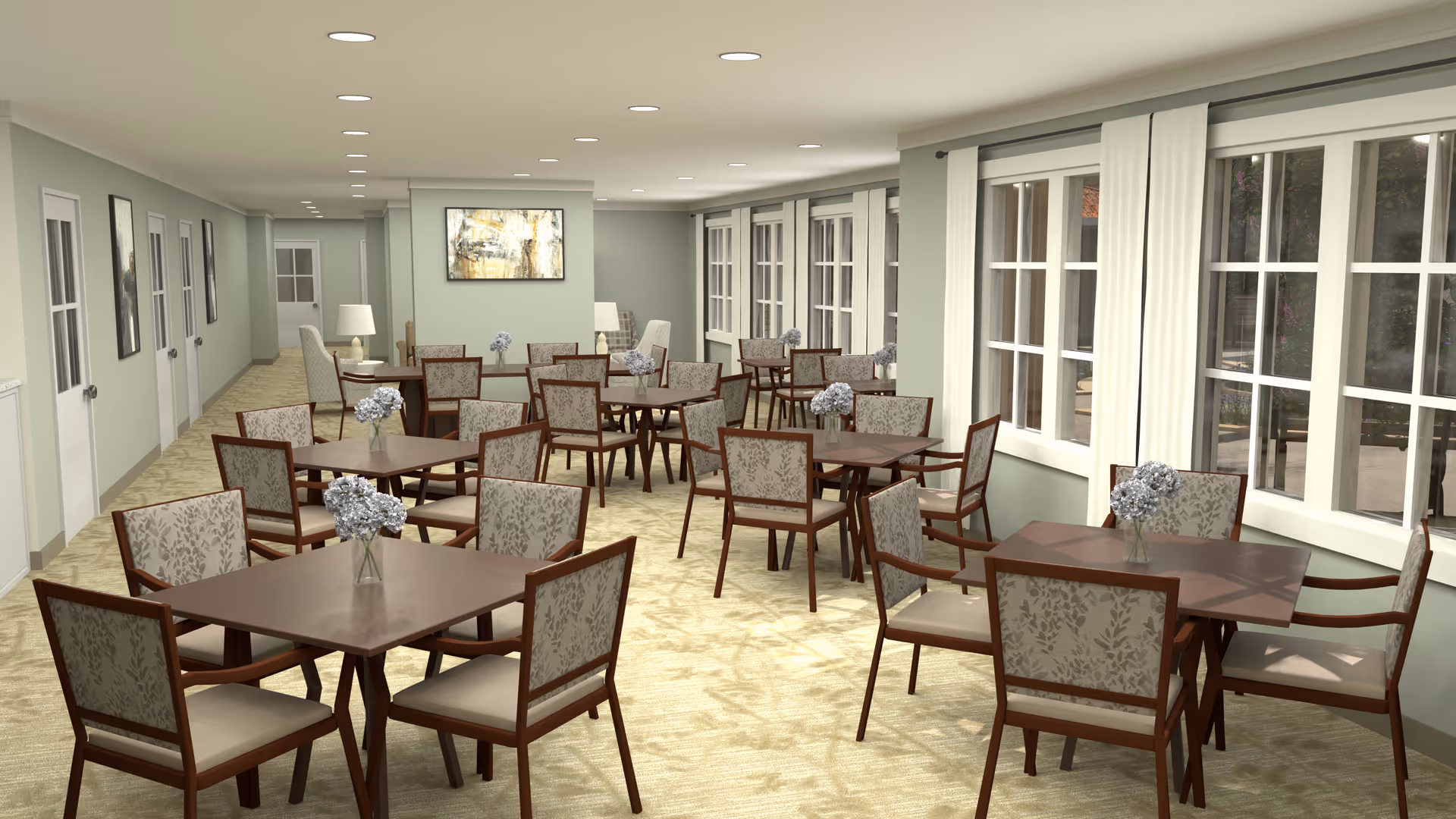 A spacious dining room with multiple square wooden tables, each surrounded by four upholstered chairs with floral patterns. Each table has a small vase with light purple flowers. The room has large windows with white curtains on the right side, light green walls, beige carpeted floor, and recessed ceiling lights. There are framed artworks and lamps along the left wall.