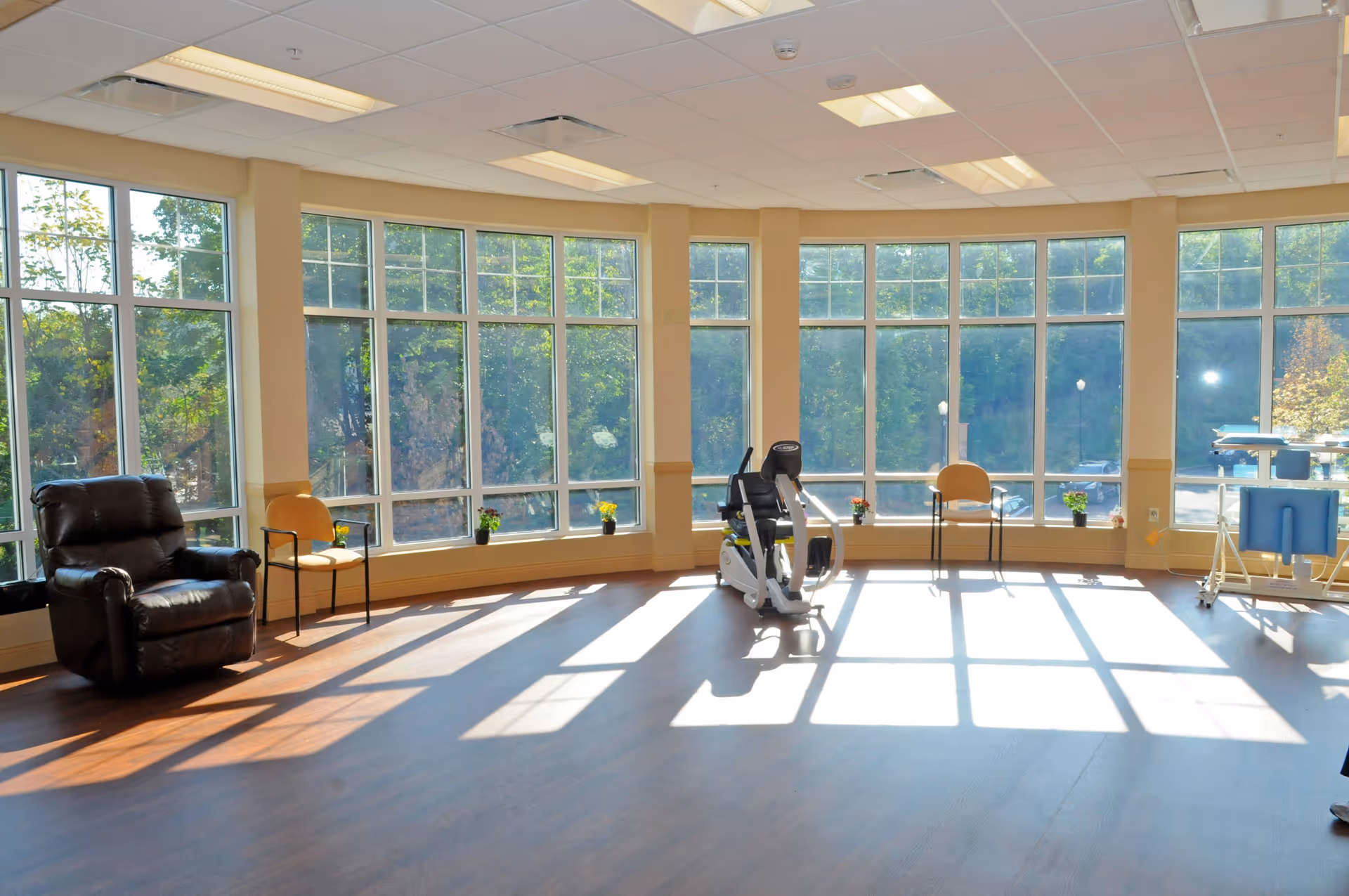A bright room with large floor-to-ceiling windows letting in natural light. The room has a dark brown recliner chair, two simple chairs, a stationary exercise bike, and some small potted plants on the window ledge. The floor is wooden, and the walls are painted beige with a white ceiling featuring recessed lighting.