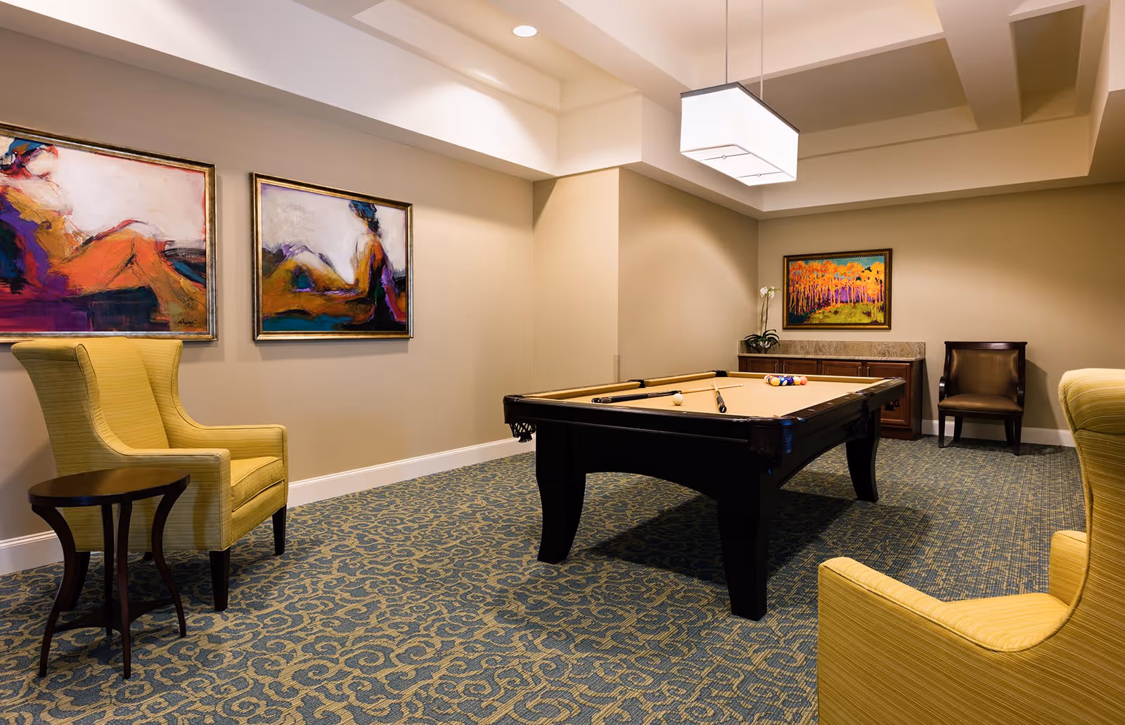 A well-lit game room with a pool table in the center, two yellow armchairs with a small wooden side table, a brown chair, and colorful abstract paintings on the beige walls. The room has a patterned carpet and a modern rectangular ceiling light.