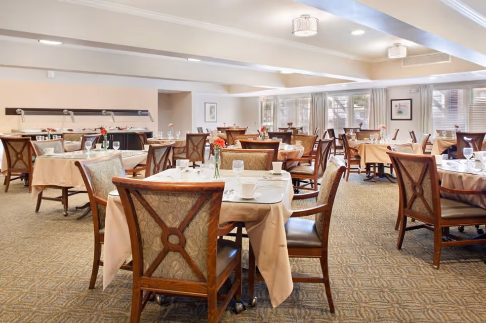 A spacious dining room with multiple tables covered in beige tablecloths, each set with white cups, glasses, and silverware. The chairs have wooden frames with patterned upholstery. The room is well-lit with ceiling lights and natural light coming through large windows with curtains. There are small flower vases on each table.