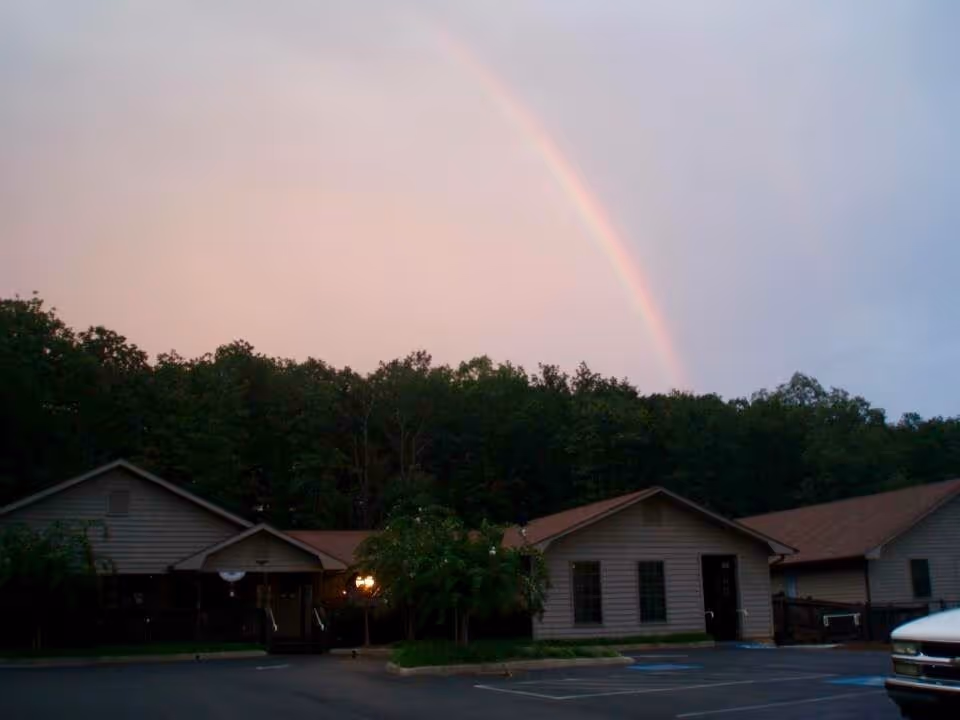 Exterior view of a single-story building with beige siding and brown roofs, surrounded by trees under a sky with a visible rainbow. The building appears to be part of a facility with a parking lot in front, including marked handicap parking spaces.