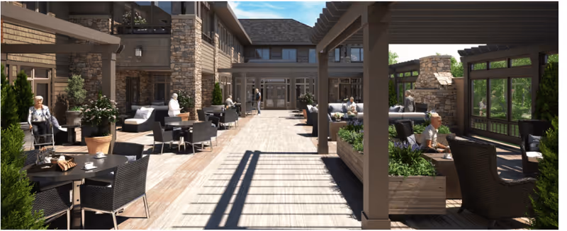 Outdoor patio area at The Kensington Reston with several elderly people sitting and relaxing on wicker chairs and sofas around tables. The space features wooden decking, potted plants, a stone fireplace, and a pergola providing partial shade. The building exterior is visible with stone and wood accents under a clear blue sky.