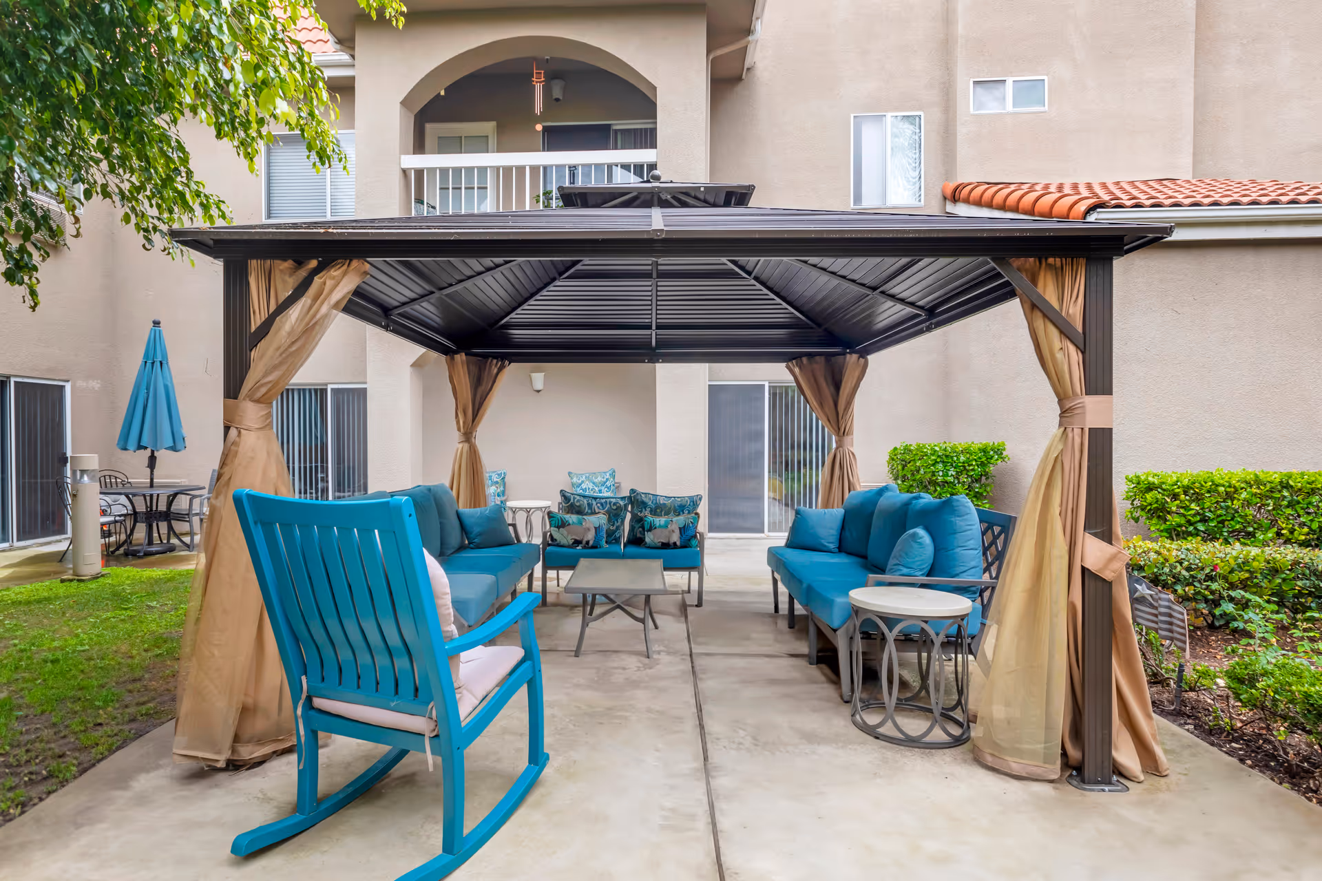 Covered outdoor patio with a gazebo, teal cushioned seating and a rocking chair in front of an apartment building.