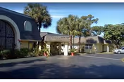 Exterior view of Pine Trail Nursing and Rehab Center showing a single-story building with a covered entrance, palm trees, and a parking area under a clear blue sky.